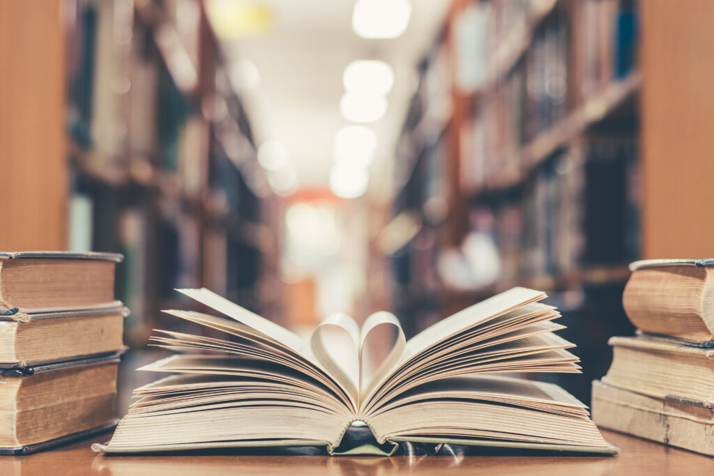 Love story book with open page of literature in heart shape and stack piles of textbooks on reading desk in library study room for education learning, and national library lovers month concept