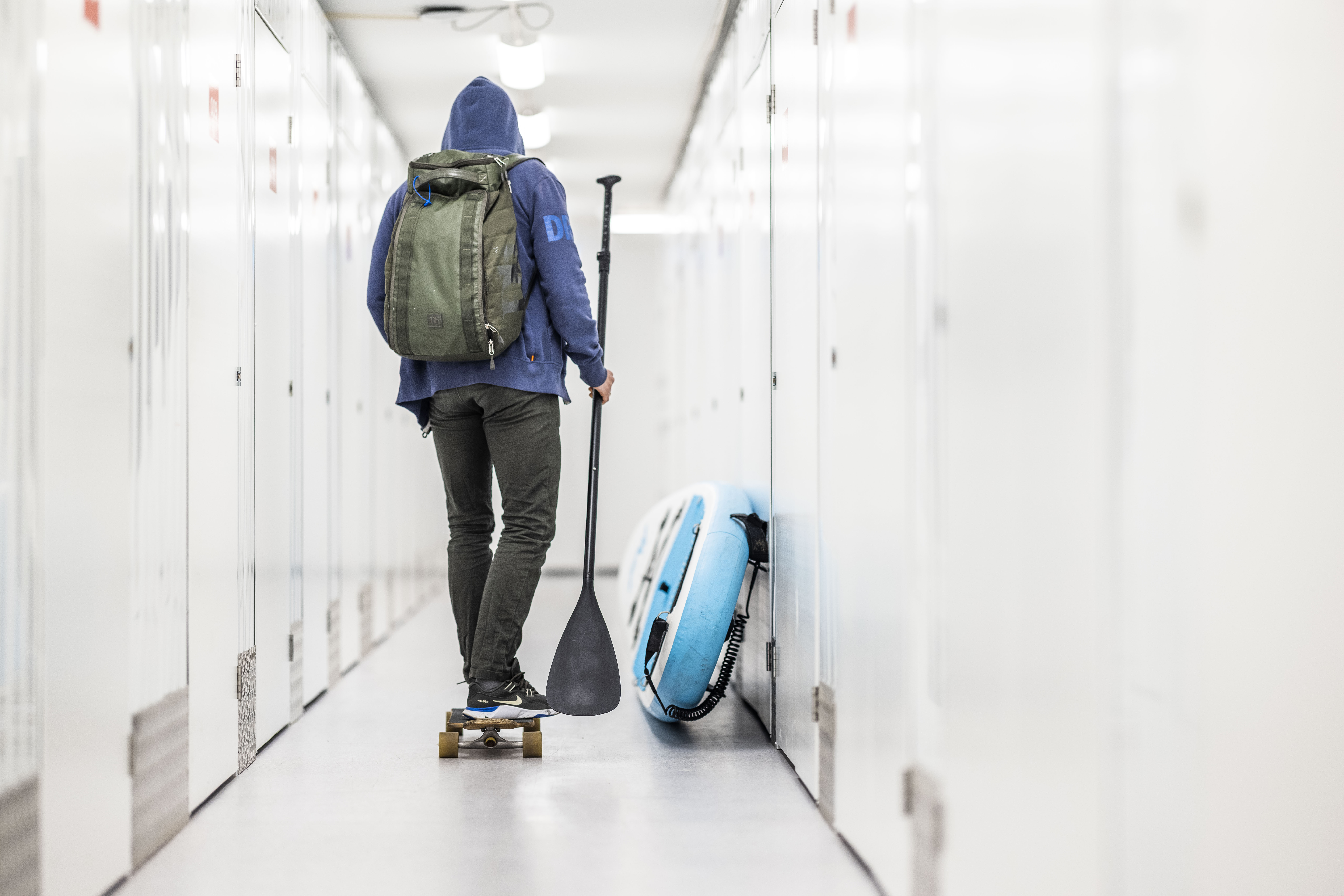 Person on a skateboard with a paddle and a backpack in a brightly lit flexistore self-storage facility, next to an inflatable paddleboard.